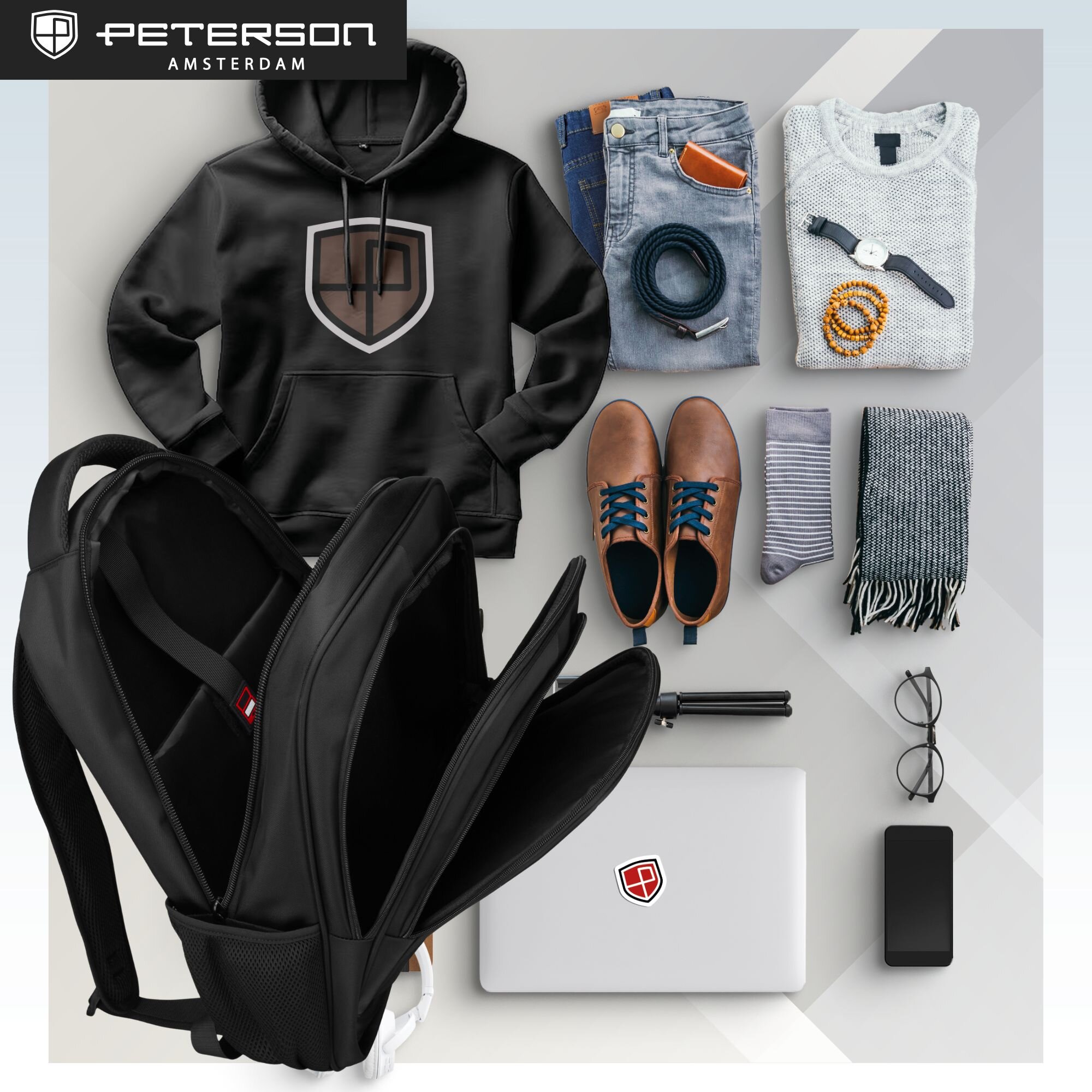 Na obrazie widoczny jest czarny plecak z kilkoma otwartymi komorami, obok którego leżą różne przedmioty, takie jak bluza z kapturem, jeansy, buty, sweter, szalik, skarpetki, laptop, okulary i telefon. W lewym górnym rogu znajduje się logo z napisem \'PETERSON AMSTERDAM\'.