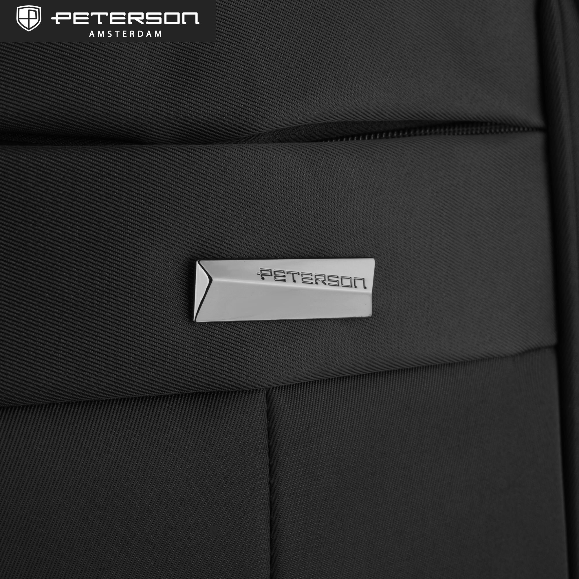 Czarny plecak z metalowym logo \'PETERSON\' na przedniej kieszeni. W górnym rogu widoczne jest logo i napis \'PETERSON AMSTERDAM\'.