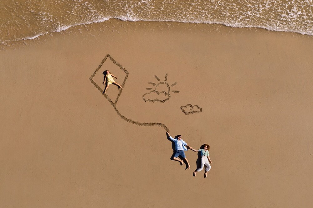 Na plaży widać trzy osoby leżące na piasku, obok których narysowany jest latawiec, słońce i chmury. W tle widoczna jest linia brzegowa z falami.
