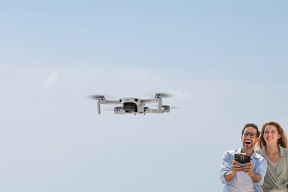 Na tle jasnego nieba widoczny jest dron w locie. Obok drona znajdują się dwie osoby, z których jedna trzyma kontroler.