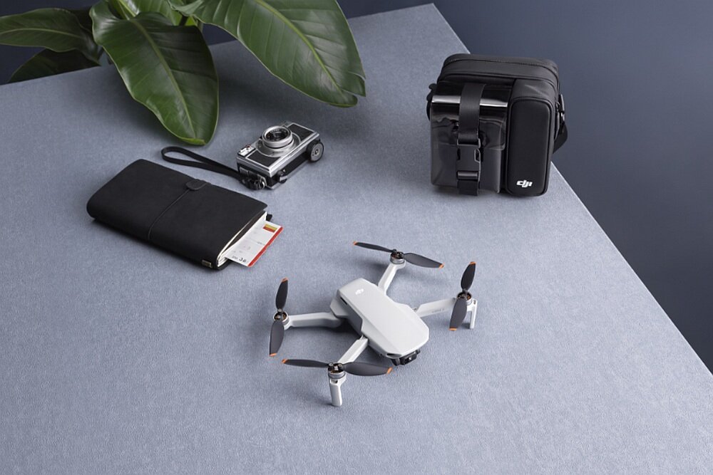 Na stole znajduje się biały dron z czterema śmigłami, obok czarna torba z logo DJI, aparat fotograficzny oraz czarny notes. W tle widoczne są zielone liście rośliny.