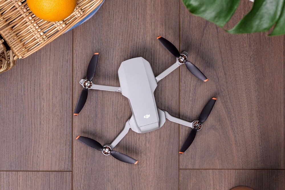 Na drewnianej powierzchni leży szary dron z czterema śmigłami, na którym widoczne jest logo DJI. Obok znajduje się kosz z pomarańczą oraz zielony liść rośliny.