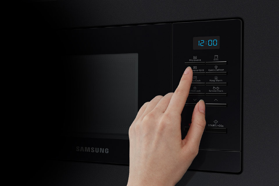 Osoba naciska przycisk na panelu sterowania kuchenki mikrofalowej z wyświetlaczem pokazującym godzinę 12:00. Na panelu widoczne są przyciski z funkcjami takimi jak Microwave, Grill, Quick Defrost oraz logo SAMSUNG.