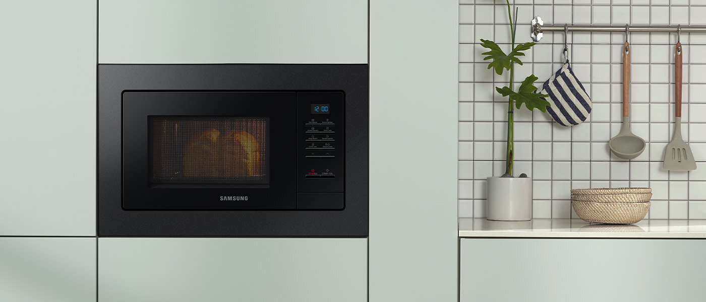 Czarna kuchenka mikrofalowa Samsung wbudowana w jasną szafkę kuchenną. Obok znajduje się ściana z białych płytek z zawieszonymi przyborami kuchennymi i rośliną w doniczce.
