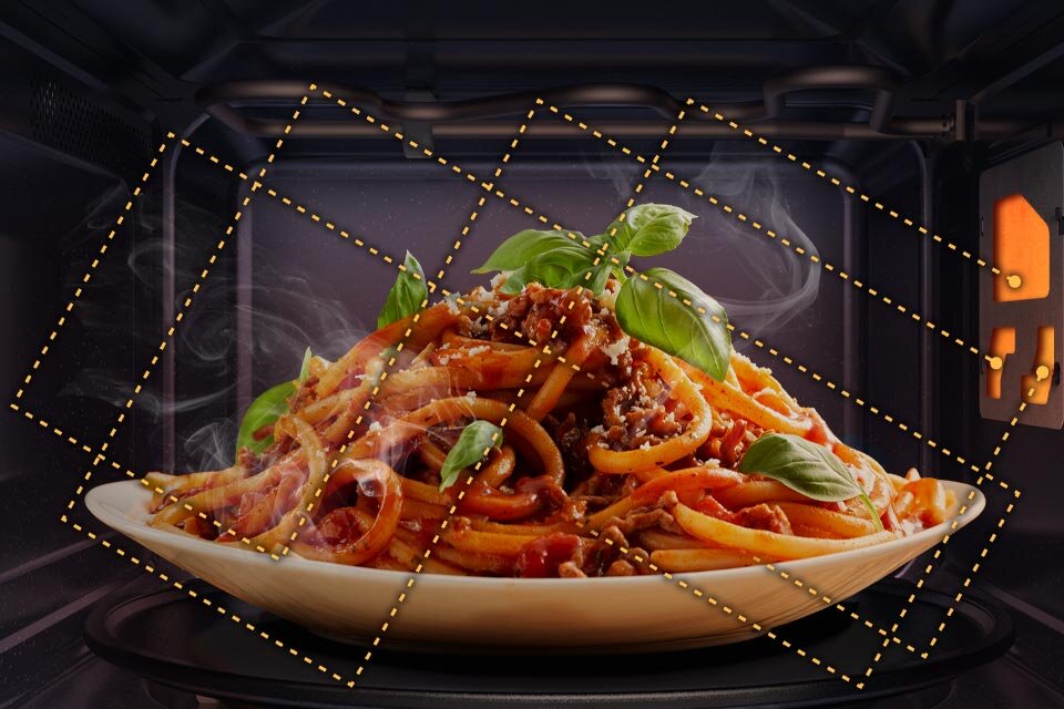 Wnętrze kuchenki mikrofalowej z talerzem spaghetti udekorowanym liśćmi bazylii. W tle widoczne są linie przerywane sugerujące równomierne podgrzewanie potrawy.
