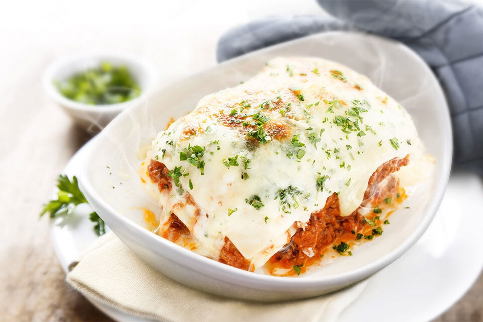 Na białym talerzu znajduje się porcja lasagne z widocznym serem i posypką z ziół. W tle widać szarą rękawicę kuchenną i niewielką miseczkę z zielonymi ziołami.