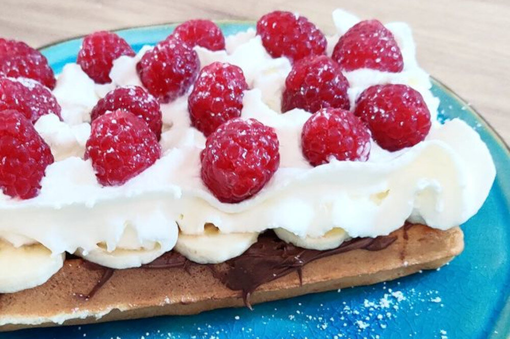 Na talerzu znajduje się gofr z bitą śmietaną, plasterkami banana, malinami i czekoladą.