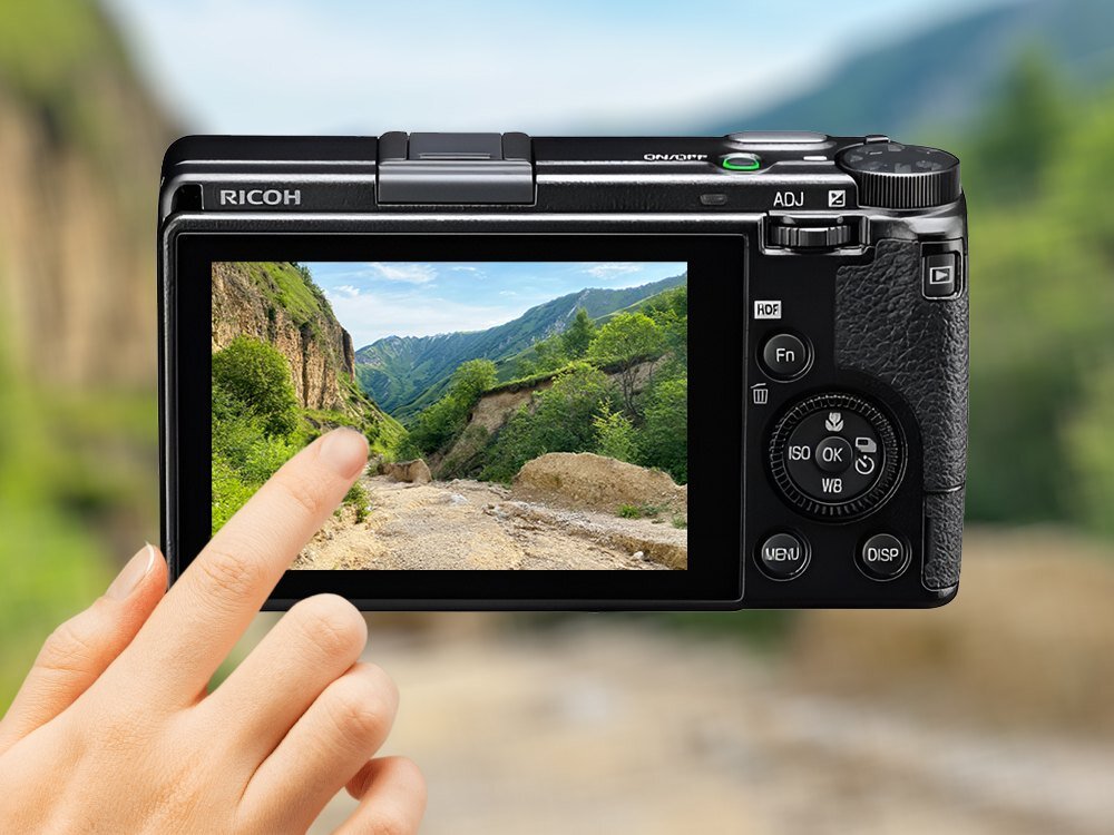 Aparat fotograficzny z widocznym ekranem LCD, na którym wyświetlany jest krajobraz górski. Na obudowie aparatu widoczne jest logo \'RICOH\'.