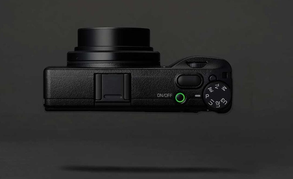 Czarny aparat fotograficzny z widocznym obiektywem i przyciskiem z napisem \'ON/OFF\'. Obok znajduje się pokrętło z oznaczeniami trybów fotograficznych.
