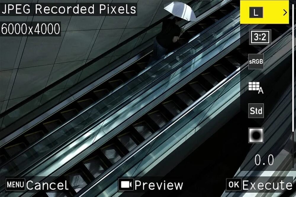 Zdjęcie przedstawia ekran aparatu z ustawieniami JPEG Recorded Pixels 6000x4000. W tle widać osobę z parasolem na ruchomych schodach.