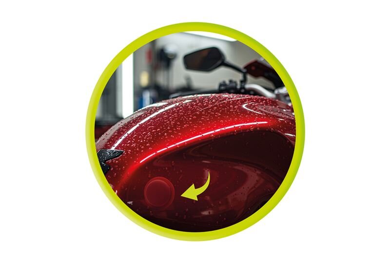 Czerwony motocykl pokryty kroplami wody z widocznym okrągłym elementem i żółtą strzałką wskazującą na niego. Obraz jest otoczony zieloną ramką.