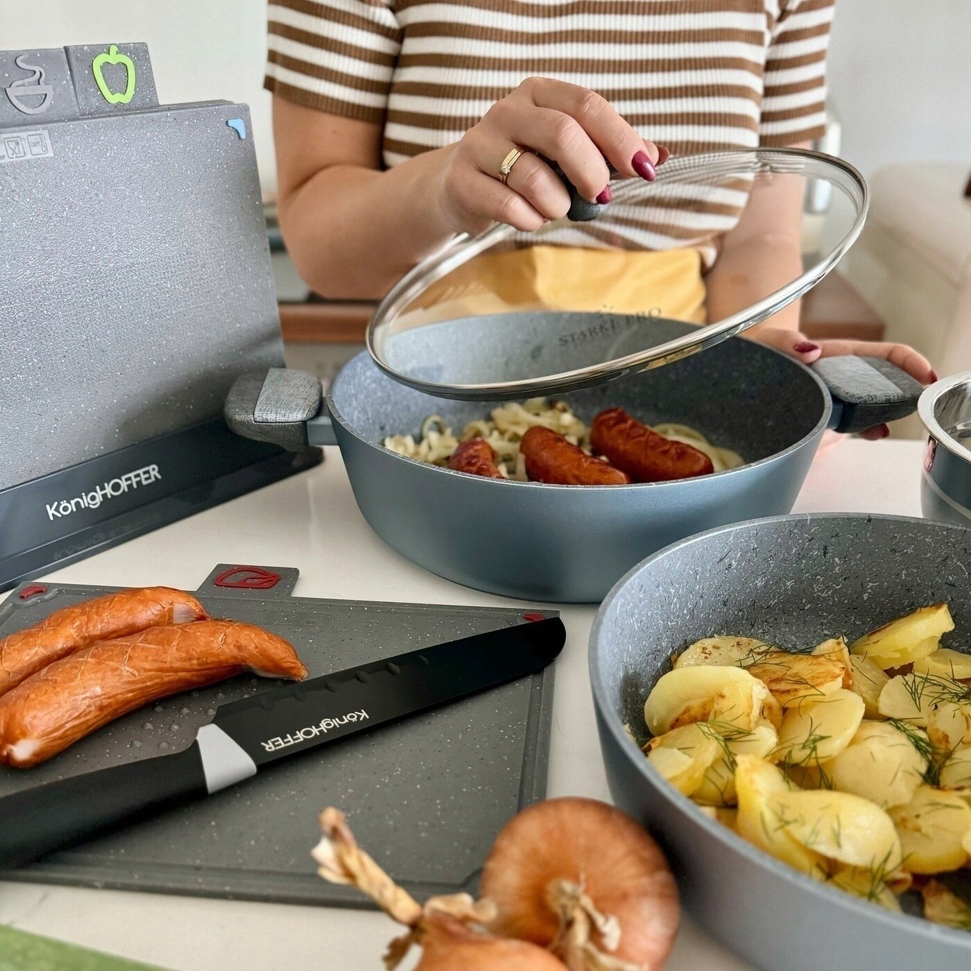 Na obrazie widać osobę trzymającą pokrywkę nad patelnią z kiełbasą i makaronem. Obok znajdują się inne naczynia z jedzeniem oraz nóż i deska do krojenia z logo KönigHOFFER.