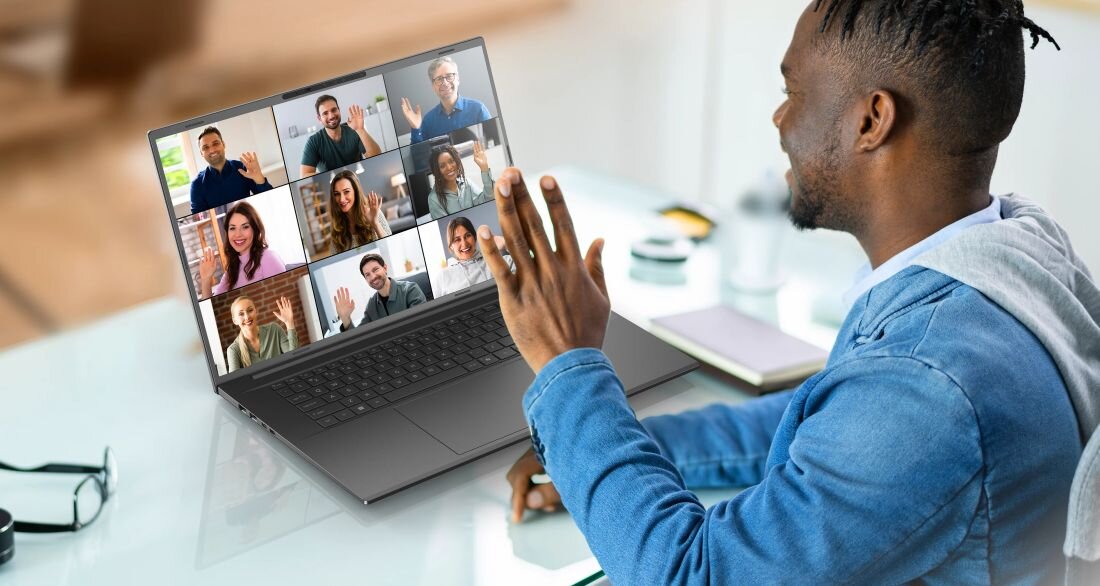 Osoba siedzi przy biurku i używa laptopa, na ekranie którego widać wideokonferencję z wieloma uczestnikami. Laptop ma duży ekran i klawiaturę, a na biurku leży długopis.