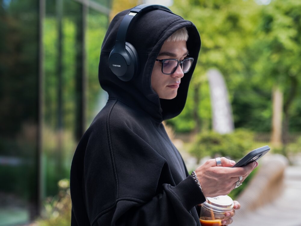 Osoba w czarnej bluzie z kapturem nosi czarne słuchawki nauszne z logo \'CANYON\' i trzyma telefon oraz napój. W tle widoczna jest zielona roślinność i szklana ściana budynku.
