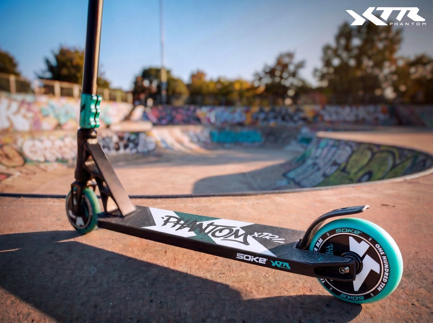 Czarna hulajnoga wyczynowa z turkusowymi kołami stoi na betonowej powierzchni skateparku. Na podeście widoczny jest napis \'PHANTOM XTR\', a w tle znajdują się graffiti na murach.