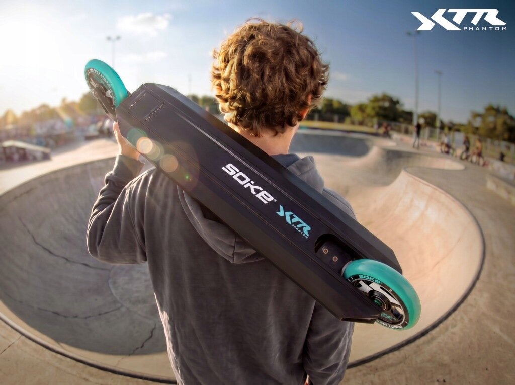 Osoba trzyma na ramieniu czarno-turkusową hulajnogę wyczynową z widocznym napisem \'SOKE XTR\'. W tle znajduje się skatepark z rampami i kilkoma osobami.