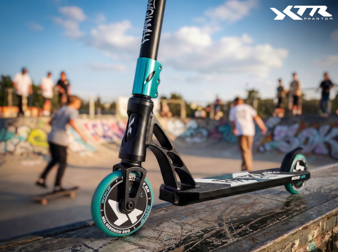 Czarna hulajnoga wyczynowa z turkusowymi elementami stoi na rampie w skateparku. W tle widać osoby korzystające z rampy, a w prawym górnym rogu znajduje się logo \'XTR PHANTOM\'.