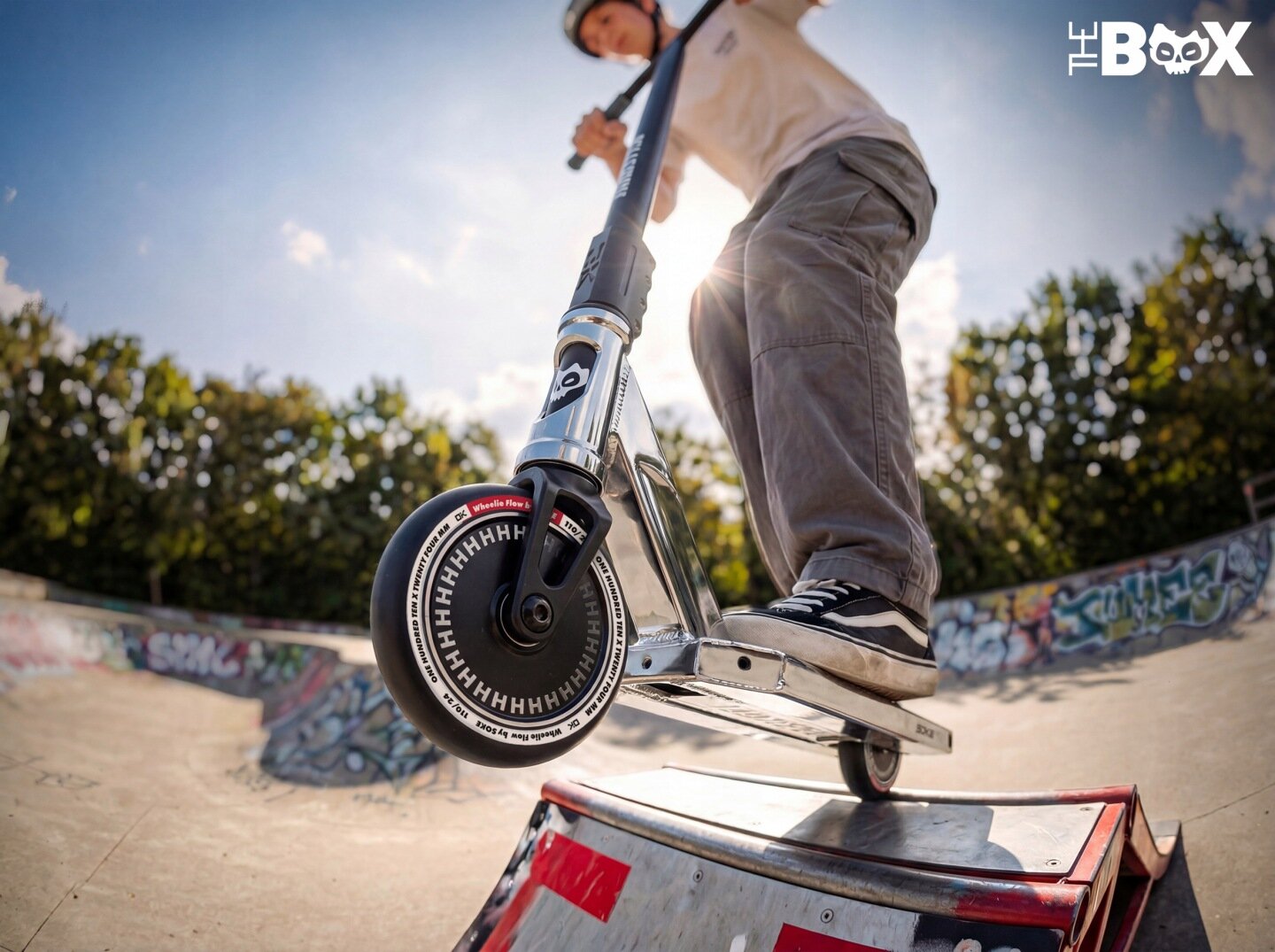 Osoba na hulajnodze wyczynowej wykonuje trik na rampie w skateparku. W tle widać drzewa i graffiti na ścianach rampy.
