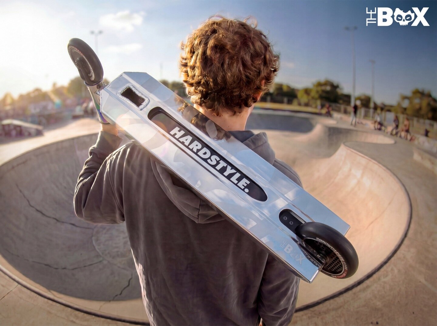 Osoba trzyma na ramieniu srebrno-czarną hulajnogę z napisem \'HARDSTYLE\'. W tle widać skatepark z betonowymi rampami.