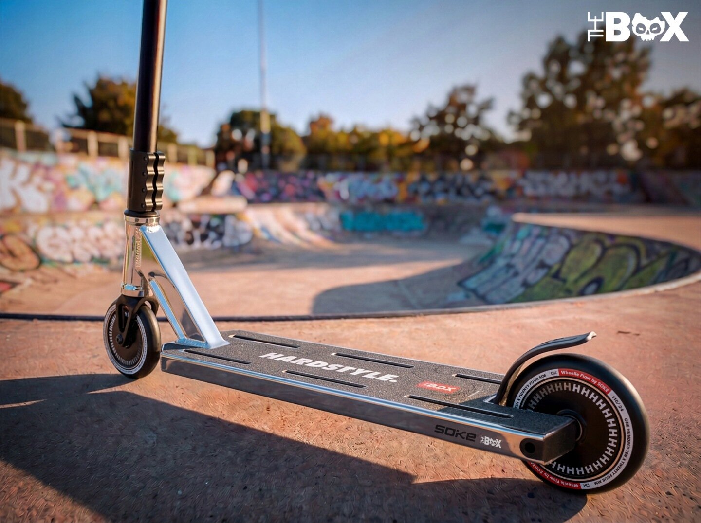 Srebrno-czarna hulajnoga wyczynowa stoi na betonowej powierzchni skateparku, w tle widać graffiti na ścianach. Na podeście hulajnogi widoczny jest napis \'HARO STYLE\', a na kole napis \'HHHHHHHHHH\'.