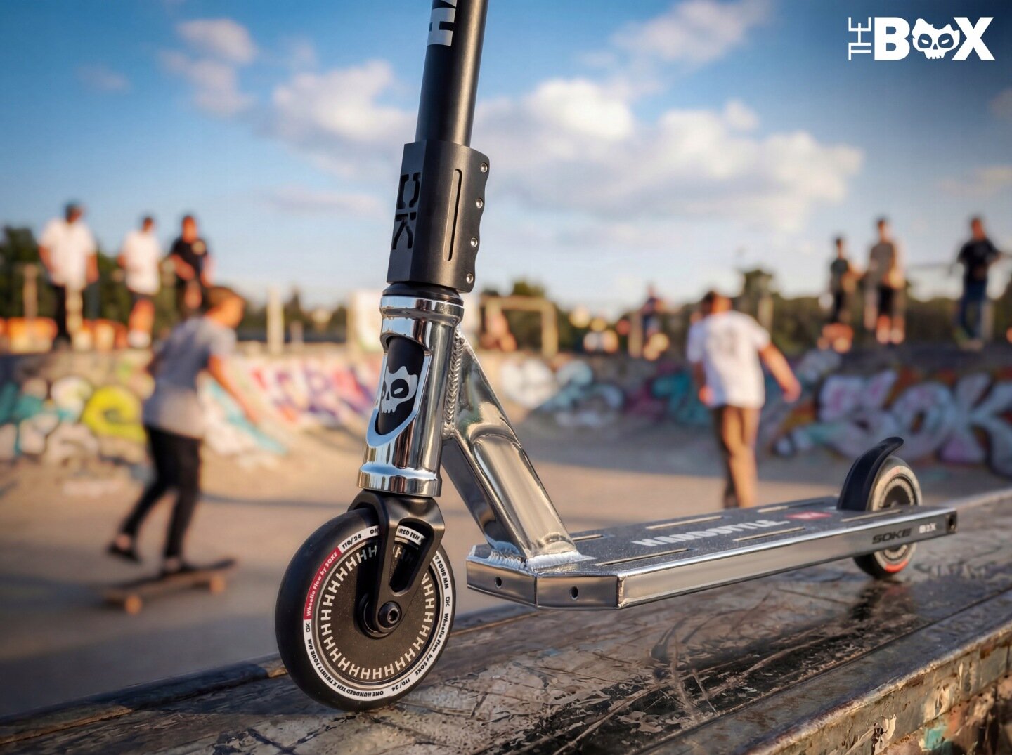 Srebrno-czarna hulajnoga wyczynowa stoi na rampie w skateparku, w tle widać osoby na deskorolkach. Na hulajnodze widoczne są napisy \'SOKE\' i \'THE BOX\'.
