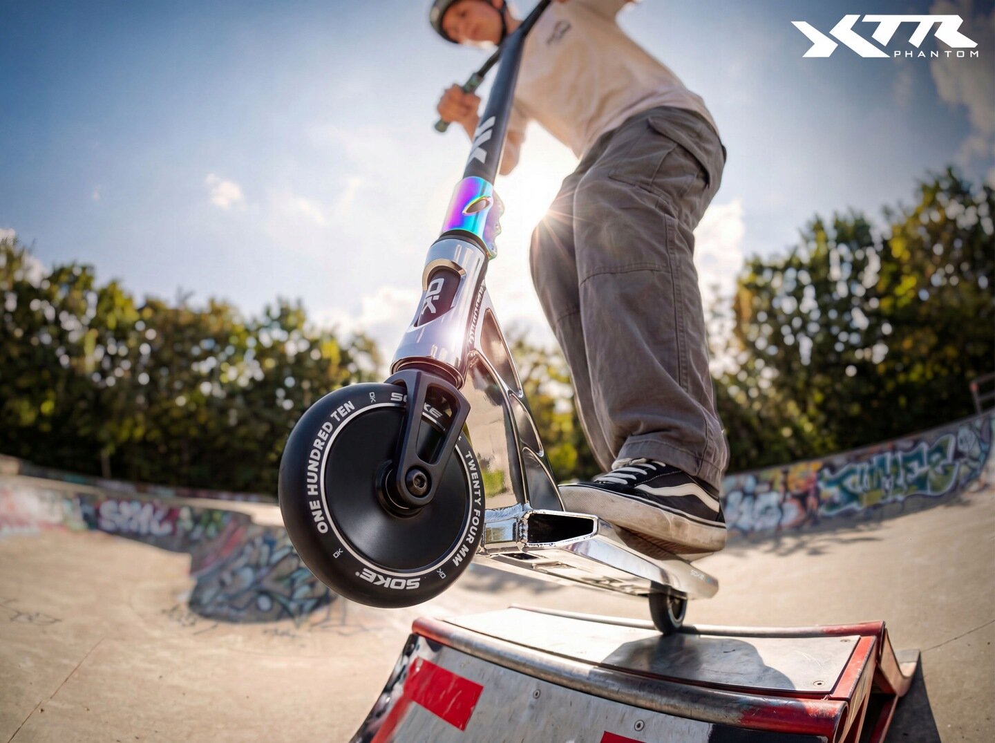 Osoba wykonuje trik na srebrno-czarnej hulajnodze wyczynowej w skateparku. Na hulajnodze widoczne są napisy \'SOKE\' i \'ONE HUNDRED TEN\'.