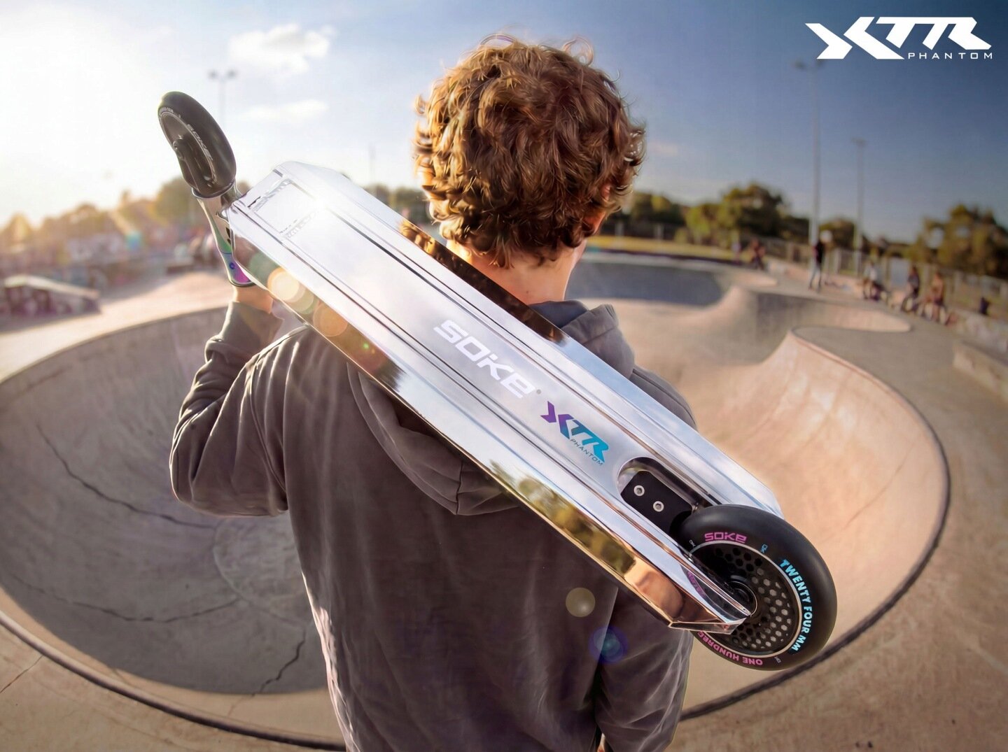 Osoba trzyma na ramieniu srebrno-czarną hulajnogę wyczynową z widocznym napisem \'SOKE XTR\'. W tle znajduje się skatepark z rampami, a w prawym górnym rogu widoczne jest logo \'XTR PHANTOM\'.