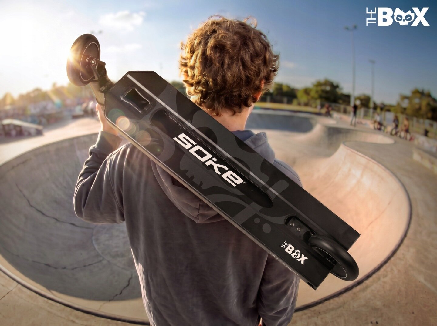 Osoba trzyma na ramieniu czarną hulajnogę z widocznym napisem \'SOKE\' oraz \'THE BOX\'. W tle znajduje się skatepark z rampami i kilkoma osobami.