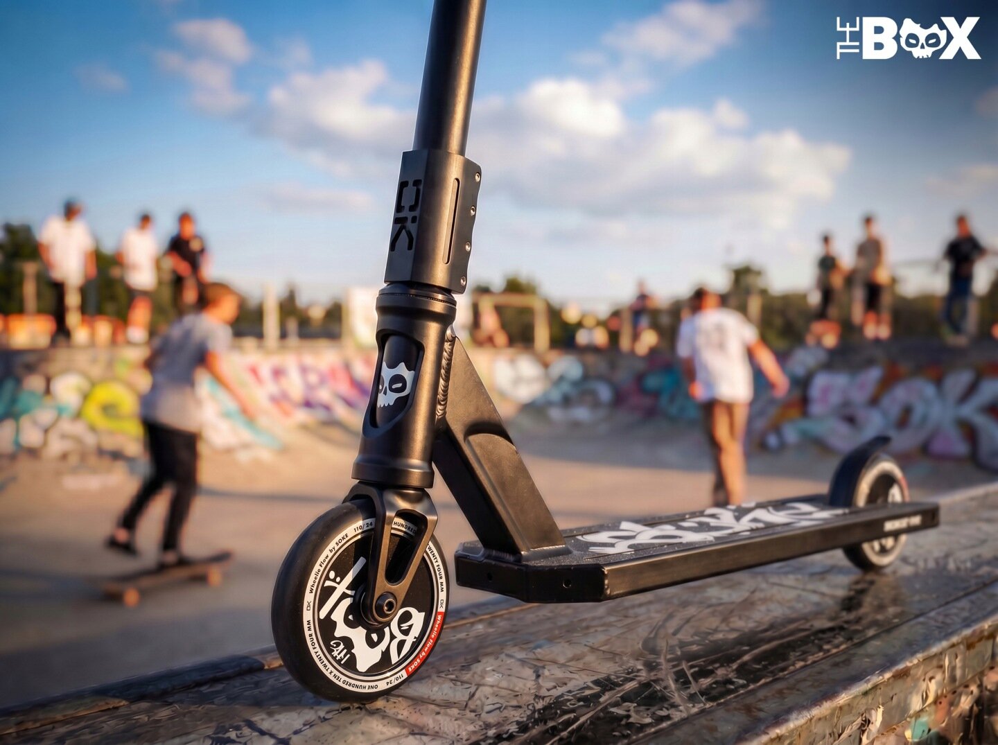 Czarna hulajnoga wyczynowa z widocznym logo czaszki na przednim widelcu stoi na rampie w skateparku. W tle widać osoby jeżdżące na deskorolkach oraz graffiti na ścianach.