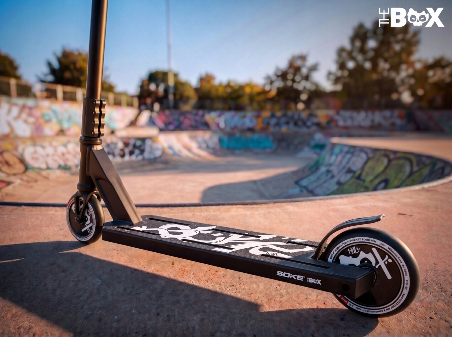 Czarna hulajnoga wyczynowa z białymi napisami na podeście stoi na tle skateparku z graffiti. Na podeście widoczny jest napis \'SOKE BOX\'.