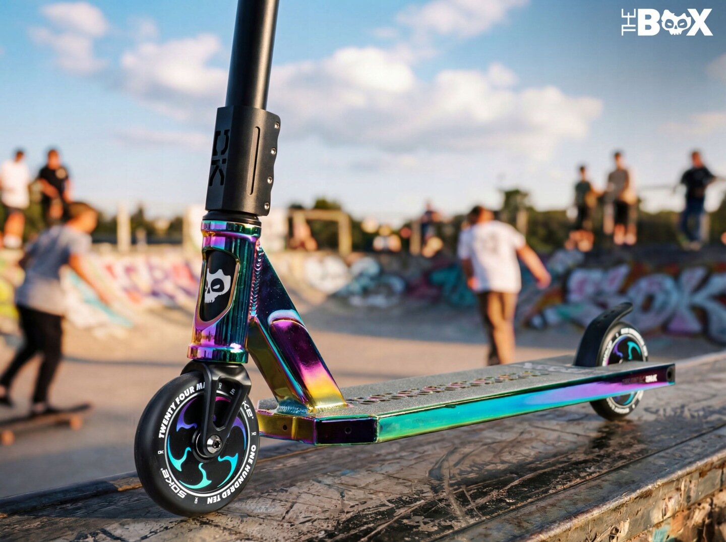 Hulajnoga wyczynowa z kolorowym, metalicznym wykończeniem stoi na rampie w skateparku. Na przednim kole widoczny jest napis \'TWENTY FOUR MM\' oraz logo \'THE BOX\' w prawym górnym rogu obrazu.