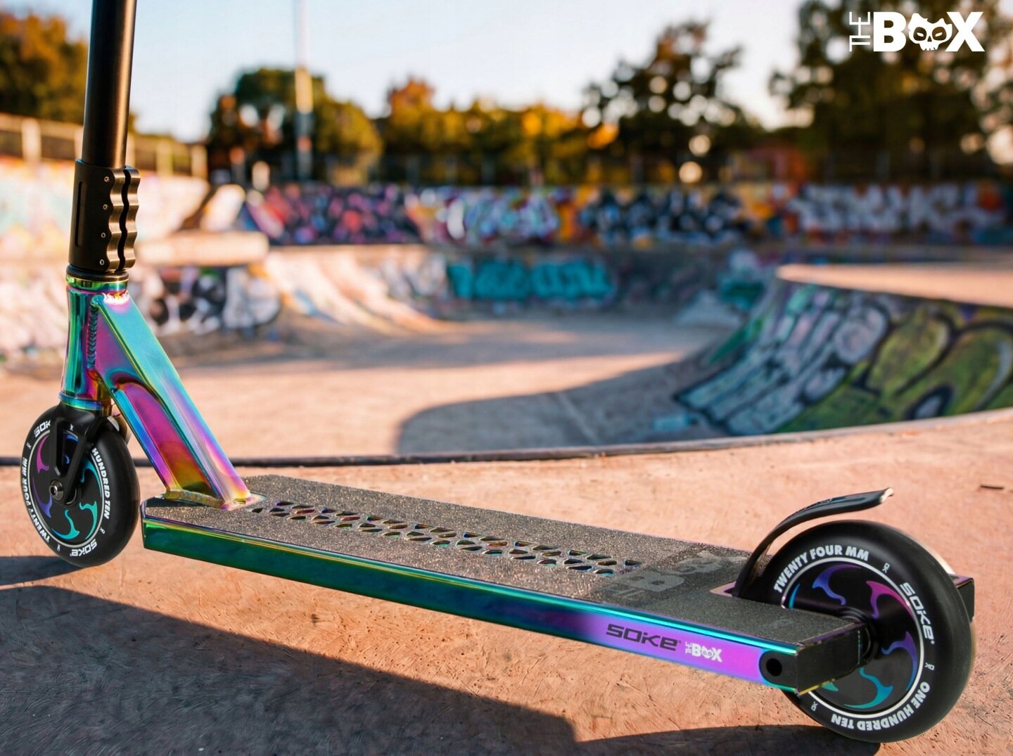 Hulajnoga wyczynowa z kolorowym, metalicznym wykończeniem stoi na tle skateparku z graffiti. Na decku widoczny jest napis \'SOKE The BOX\'.