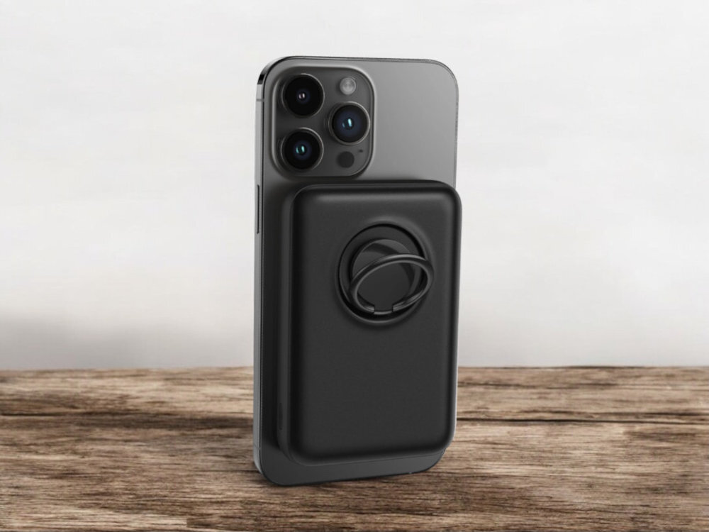 Na drewnianym blacie stoi smartfon z przymocowanym czarnym powerbankiem. Powerbank ma okrągły uchwyt na tylnej części.