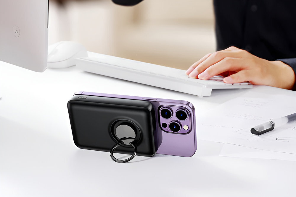 Na biurku znajduje się czarny powerbank z pierścieniem, przymocowany do fioletowego smartfona. W tle widać ręce osoby piszącej na klawiaturze oraz monitor.