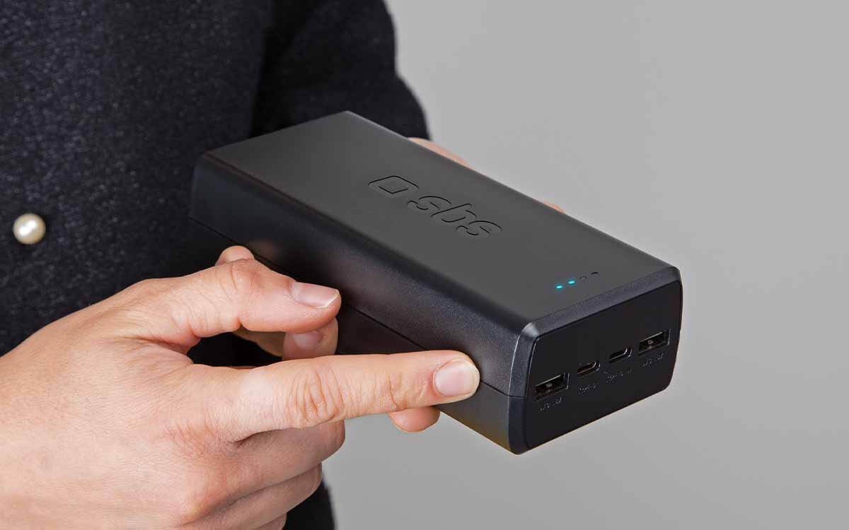 Osoba trzyma czarny powerbank z logo \'sbs\' i czterema portami na jednym z boków. Na górze urządzenia widoczne są cztery niebieskie diody LED.