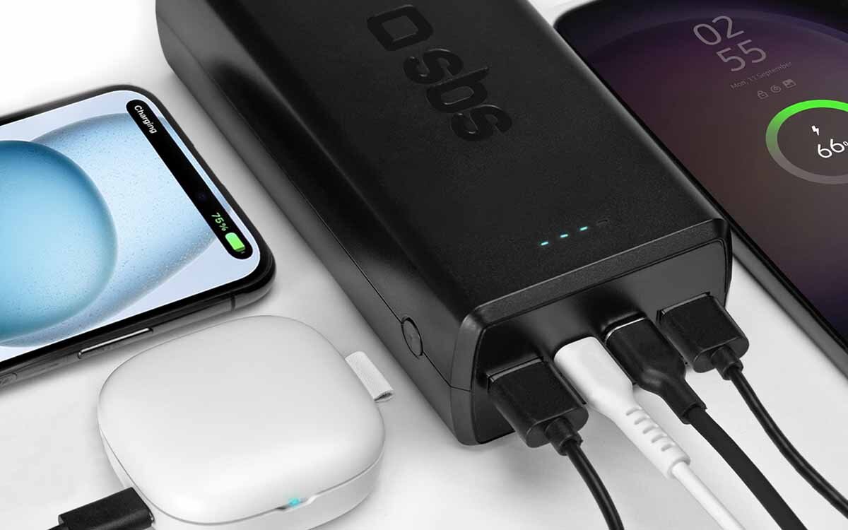 Na obrazie widoczny jest czarny powerbank z logo \'sbs\', do którego podłączone są trzy kable. Obok znajdują się dwa smartfony i białe urządzenie elektroniczne.