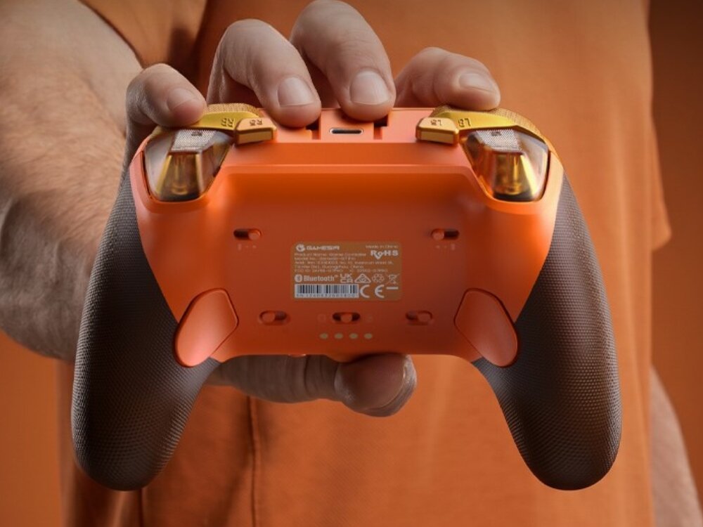 Osoba trzyma pomarańczowy kontroler z czarnymi uchwytami. Na kontrolerze widoczne są napisy: \'GAMESIR\', \'RoHS\', \'Bluetooth\'.