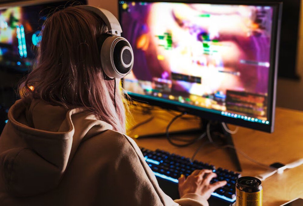 Procesor AMD Ryzen 7 7700X3D Osoba w bluzie z kapturem i słuchawkach siedzi tyłem, grając w grę na monitorze. Ręka na podświetlanej klawiaturze, obok puszka