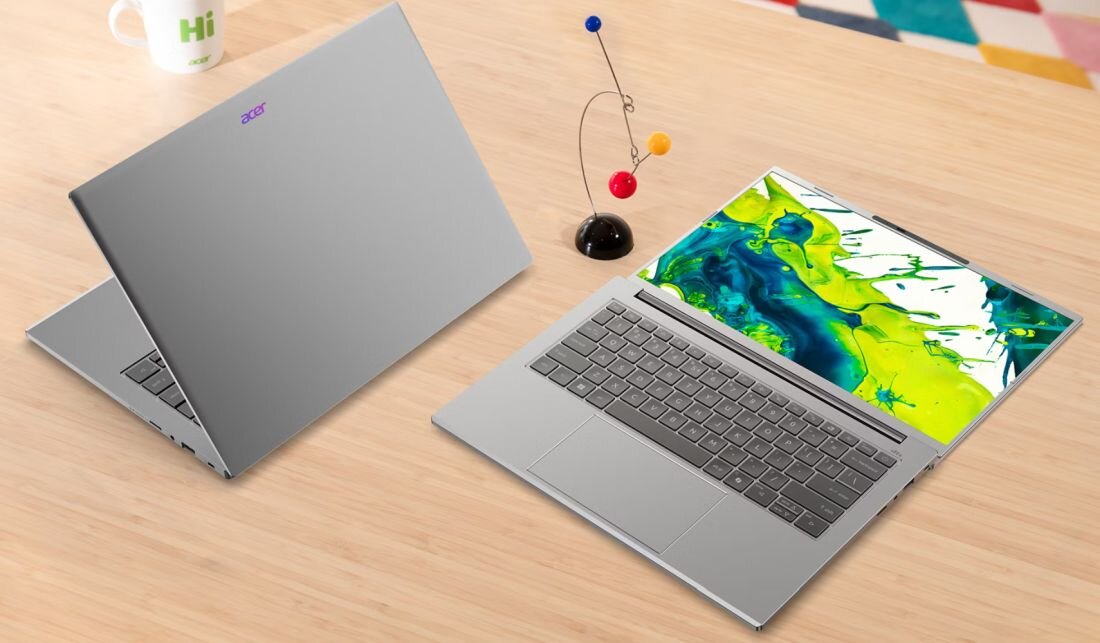 Na drewnianym stole znajdują się dwa laptopy w kolorze srebrnym, jeden otwarty z widocznym kolorowym ekranem, drugi zamknięty z widocznym logo Acer. Obok laptopów stoi dekoracyjna figurka z kulkami na drucikach oraz biały kubek z napisem \'Hi\'.