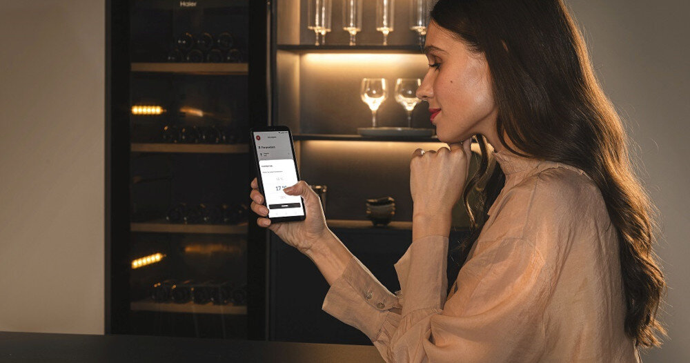 Osoba trzymająca smartfon z widocznym ekranem, na którym wyświetlane są opcje związane z temperaturą. W tle znajduje się chłodziarka do wina z widocznymi butelkami i kieliszkami.