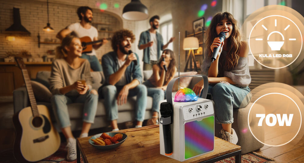 Grupa osób bawi się w salonie, jedna z nich śpiewa do mikrofonu. Na pierwszym planie widoczny jest głośnik karaoke z kolorowym oświetleniem LED i napisem \'KULA LED RGB\' oraz \'70W\'.