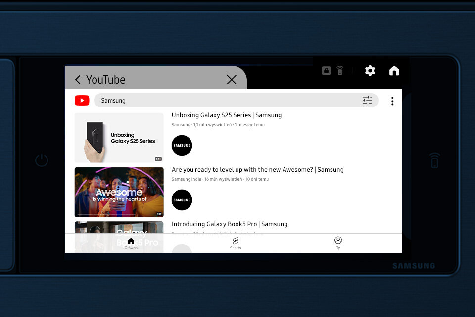 Ekran urządzenia wyświetla aplikację YouTube z wyszukiwaniem \'Samsung\'. Widoczne są trzy wyniki: \'Unboxing Galaxy S25 Series | Samsung\', \'Are you ready to level up with the new Awesome? | Samsung\', oraz \'Introducing Galaxy Book5 Pro | Samsung\'.