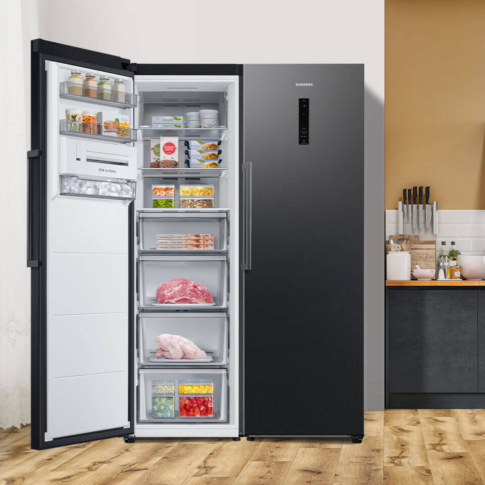 Większy zakres otwarcia w zamrażarce TWIN ułatwia korzystanie z urządzenia Otwarta zamrażarka z widocznymi półkami i szufladami wypełnionymi różnymi produktami spożywczymi. Na drzwiach zamrażarki widnieje logo \'Samsung\'.