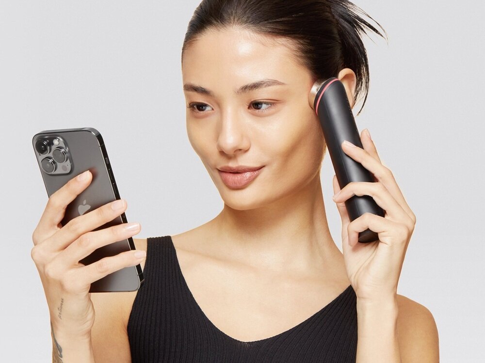 Osoba trzyma w jednej ręce czarne urządzenie do pielęgnacji twarzy, a w drugiej smartfon. Urządzenie ma metaliczną końcówkę i jest przyłożone do twarzy.