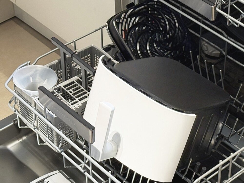 Wnętrze zmywarki z umieszczonym w niej białym urządzeniem kuchennym z czarną pokrywą i uchwytem. Obok znajduje się plastikowy lejek i metalowy koszyk na sztućce.