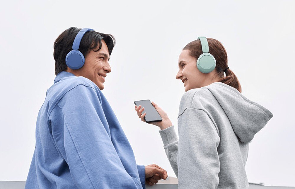 Słuchawki nauszne SONY WH-CH535 Mężczyzna i kobieta stoją obok siebie na zewnątrz, oboje mają na sobie słuchawki nauszne. Kobieta trzyma telefon i uśmiecha się do mężczyzny