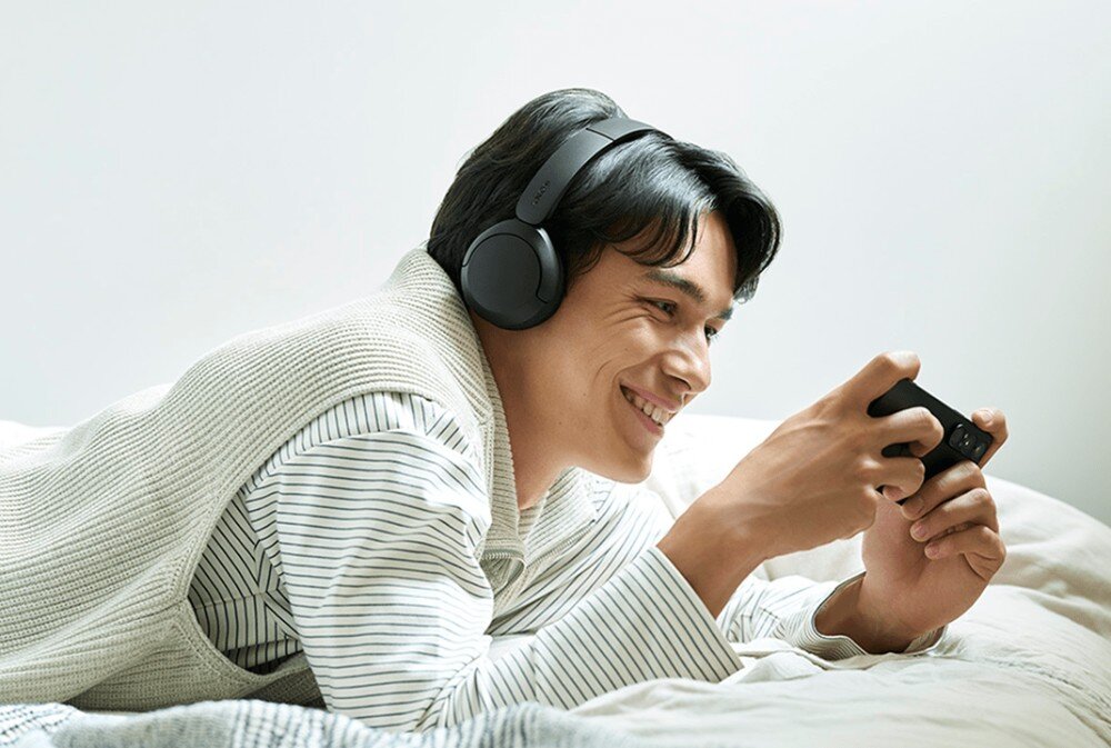 Słuchawki nauszne SONY WH-CH535 Mężczyzna leży na łóżku, trzyma w obu dłoniach telefon i uśmiecha się, mając na sobie słuchawki nauszne