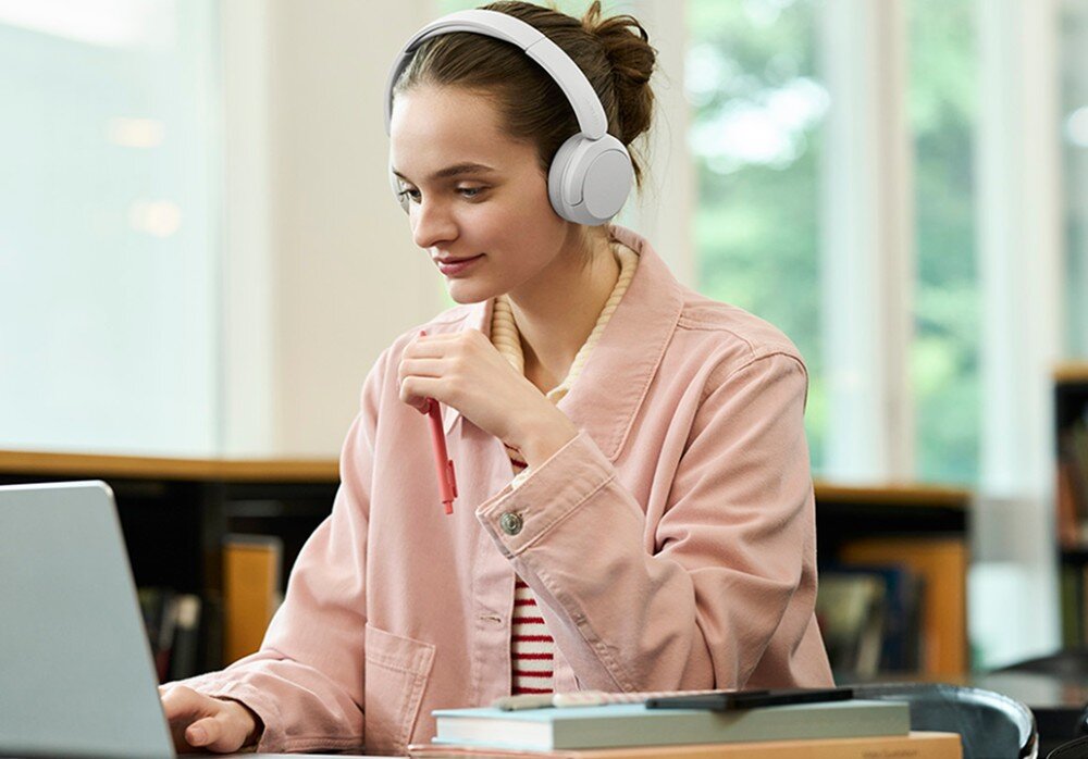Słuchawki nauszne SONY WH-CH535 Kobieta w różowej kurtce siedzi przy biurku przed otwartym laptopem, trzyma długopis w dłoni i nosi słuchawki nauszne