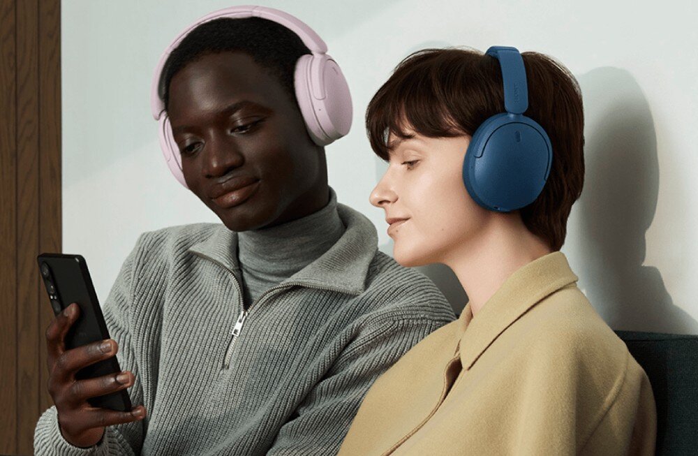 Słuchawki nauszne SONY WH-CH735N Mężczyzna i kobieta blisko siebie. Mężczyzna patrzy na telefon, kobieta na niego. Oboje mają na uszach słuchawki nagłowne, jasne i ciemnoniebieskie, na jasnym tle