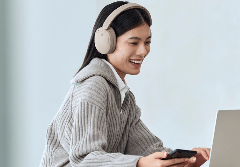 Słuchawki nauszne SONY WH-CH735N Kobieta w szarym swetrze siedzi przy laptopie, uśmiechając się. Patrzy w ekran, na uszach ma beżowe słuchawki nagłowne. W dłoni trzyma smartfon na jasnym tle